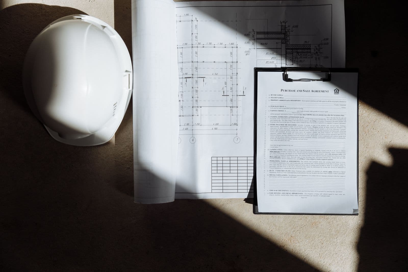 Des plans, un contrat et un casque de chantier posés au soleil.
